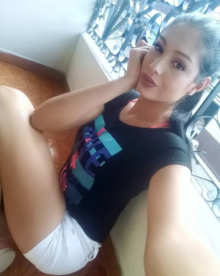 Femme assise près d'une fenêtre, en t-shirt noir et shorts blancs, main sur la joue, regardant l'appareil.