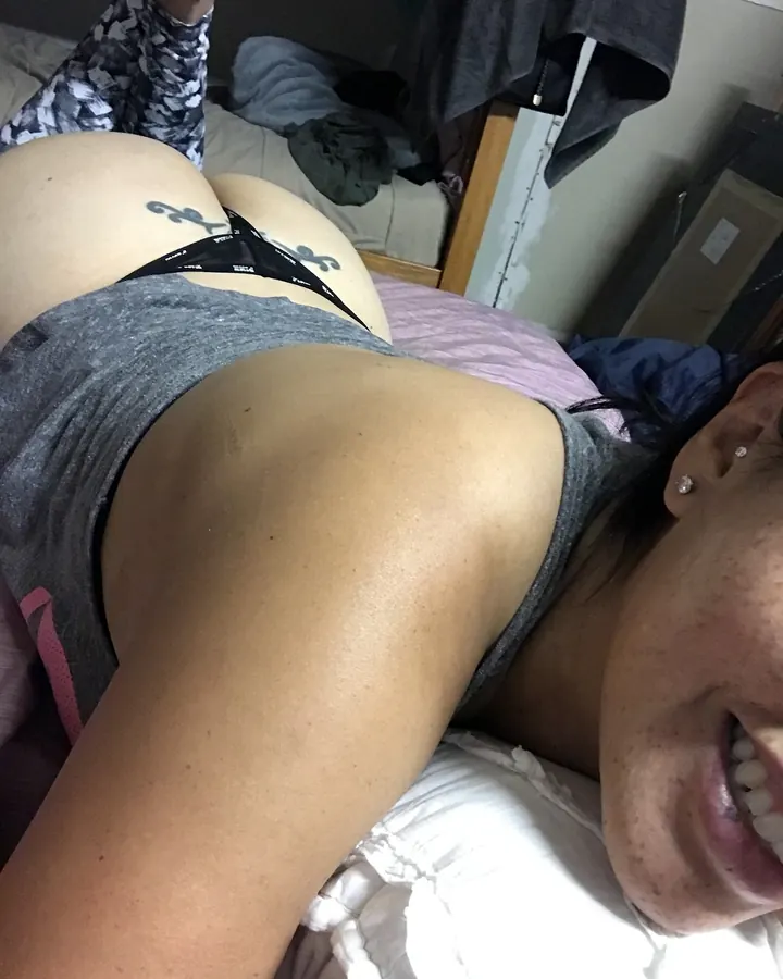 Femme allongée sur un lit, en haut gris et sous-vêtements noirs, regardant l'appareil.