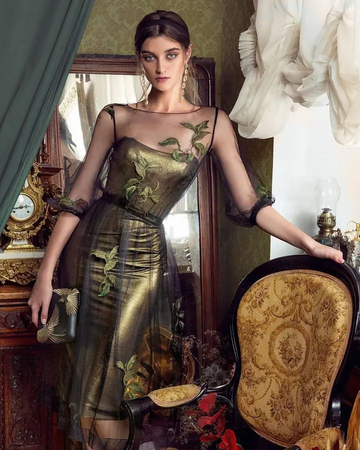 Femme en robe dorée transparente avec broderies vertes, debout près d'un miroir dans un décor ancien.