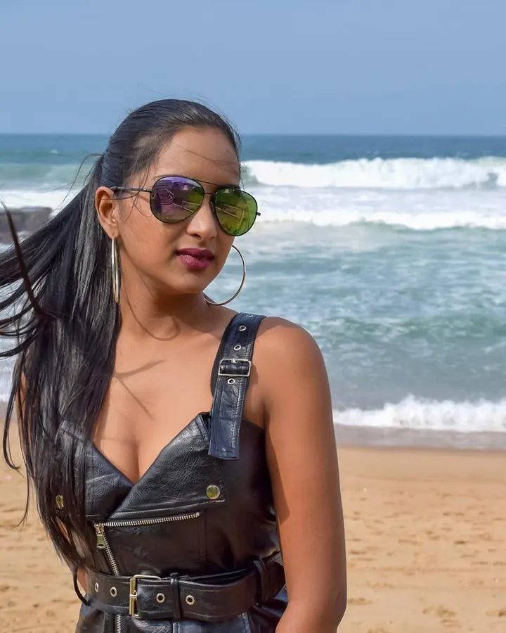 Femme en tenue de cuir noir, lunettes de soleil, debout sur une plage, vent dans les cheveux.