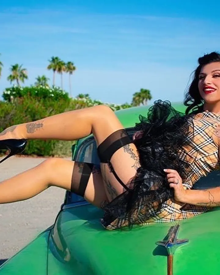 Femme arabe aux cheveux noirs et tatouages, en robe à carreaux et bas noirs, assise sur le capot d'une voiture verte sous un ciel bleu.