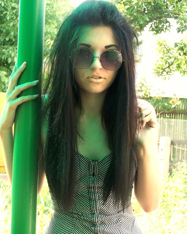 Femme arabe de 25 à 35 ans, portant des lunettes de soleil et un haut à carreaux, appuyée contre un poteau vert en extérieur.