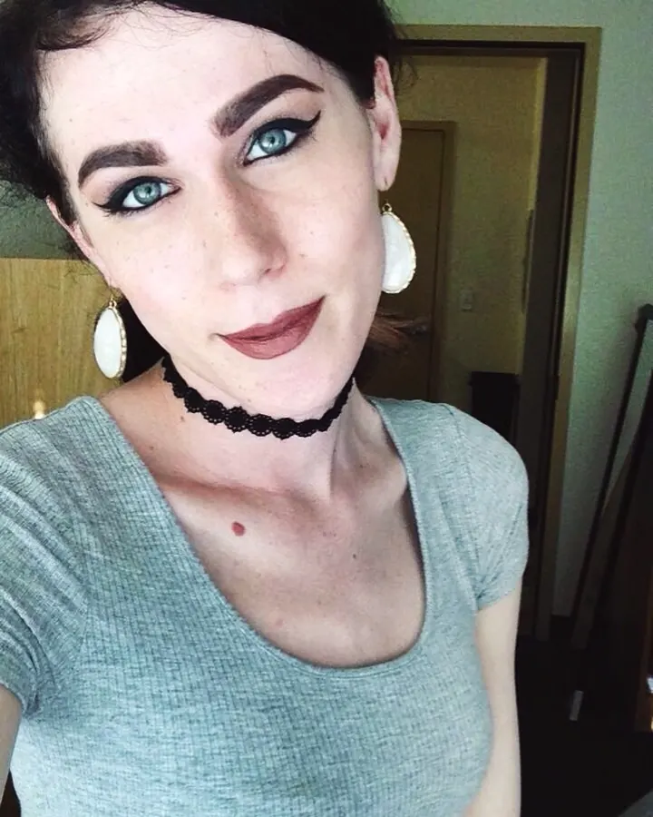 Femme arabe de 18 à 25 ans, portant un haut gris, souriante, regardant vers l'appareil, collier noir et boucles d'oreilles blanches.