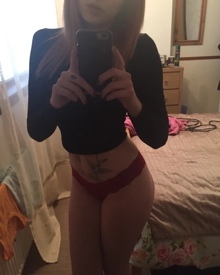Femme aux cheveux châtains et yeux gris, en haut noir et culotte rouge, prenant un selfie devant un miroir dans une chambre.