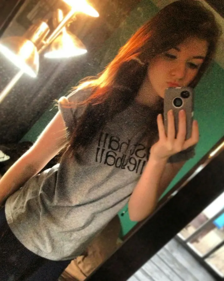 Jeune femme arabe aux cheveux roux, prenant un selfie devant un miroir avec une lampe à côté, vêtue d'un t-shirt gris.