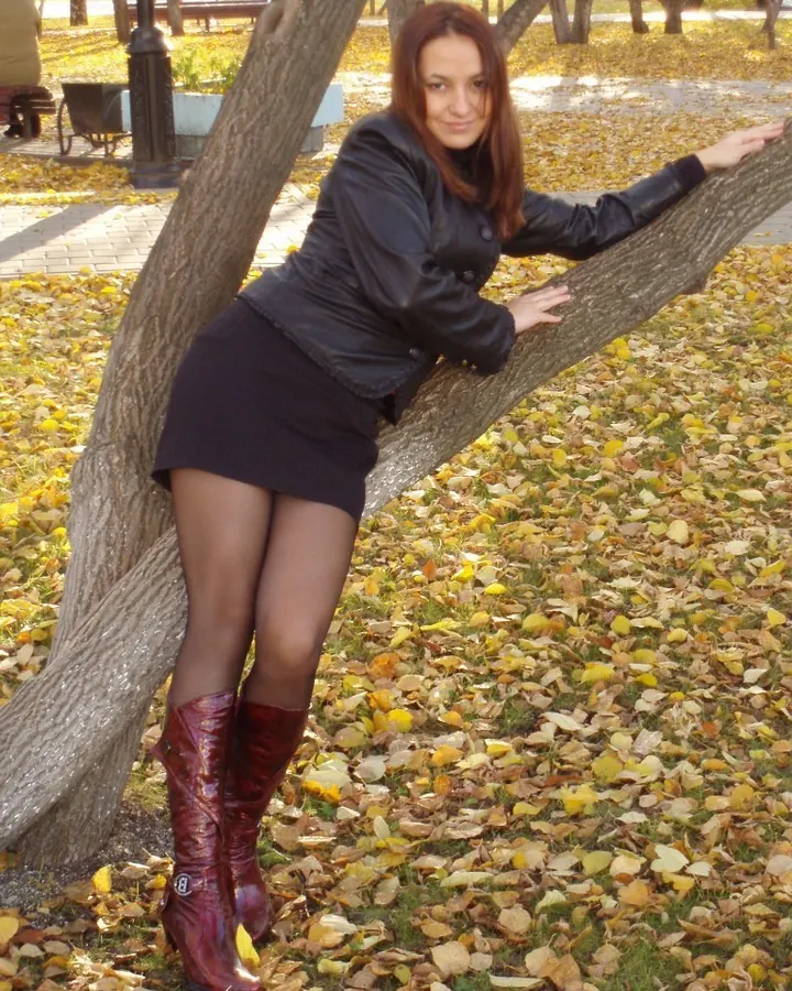 Femme arabe de 25 à 35 ans en robe noire courte, posant contre un tronc d'arbre dans un parc automnal avec des feuilles mortes.