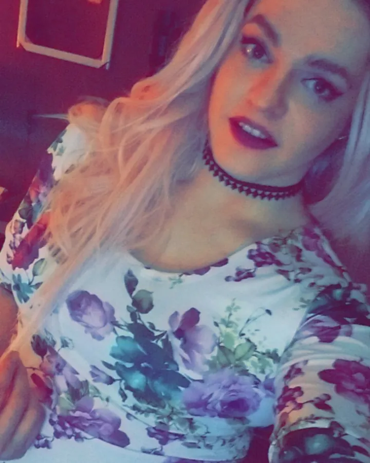 Femme aux cheveux blonds portant un haut noir, regardant directement la caméra, dans un intérieur sombre