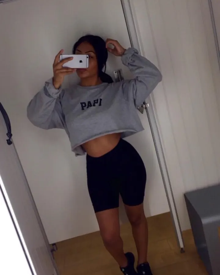 Femme en robe noire, assise sur un lit, collant noir, cheveux bruns, regardant l'appareil.