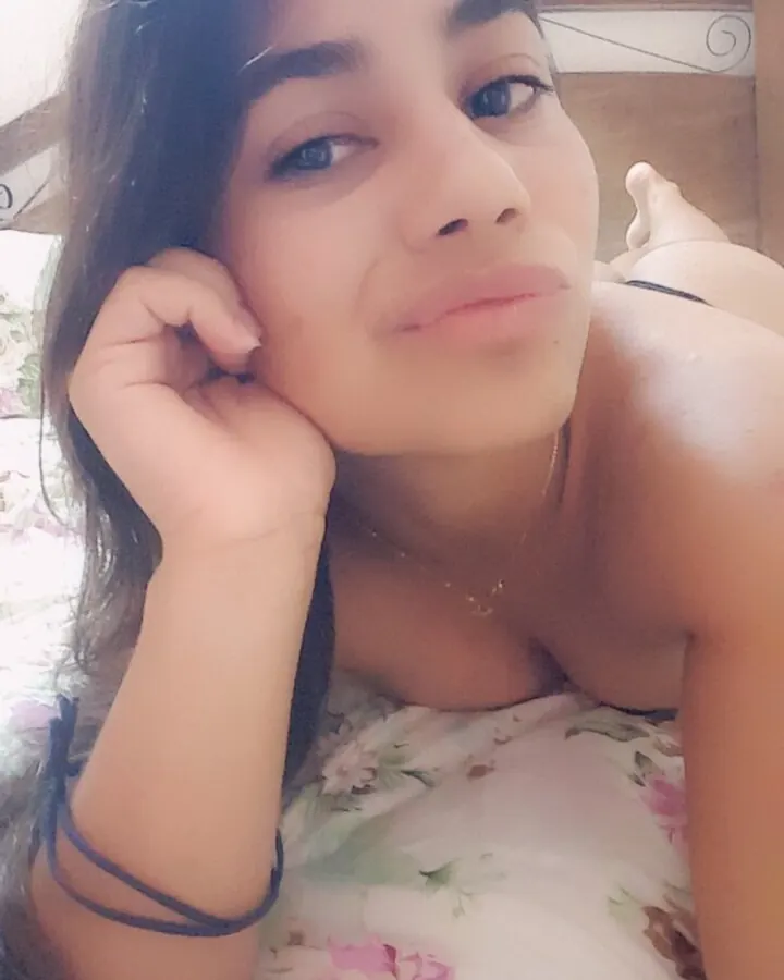 Femme souriante, cheveux foncés, regardant vers le haut, main près de l'oreille, chemise fleurie.