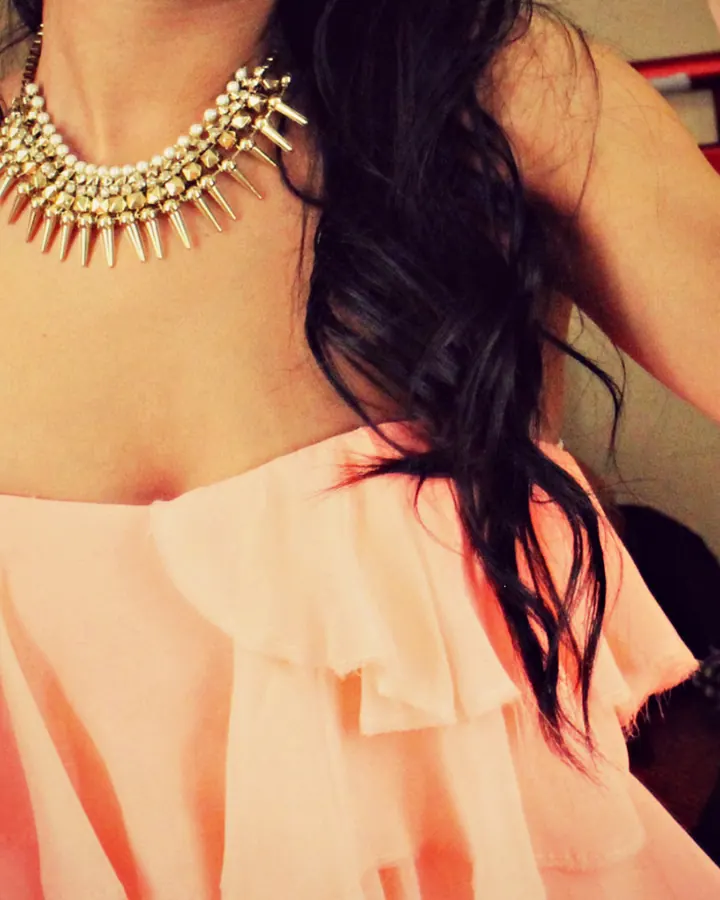 Femme en robe rose décolletée, collier doré pointu, cheveux noirs ondulés, main levée.